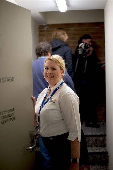 Staff Member guiding a tour group holds open the back stage door smiling at camera